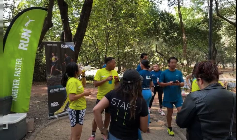 Un grupo de personas vestidas con ropa deportiva se reúne cerca de pancartas verdes en un parque, en representación de Hatch Co. Algunos sostienen bebidas mientras otros conversan. Al fondo se ven árboles y senderos, lo que se suma al ambiente vibrante de este evento de agencia de marketing.