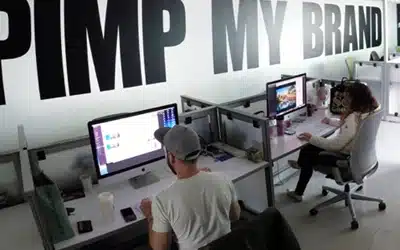 Dos personas trabajan frente a computadoras en un espacio de oficina moderno debajo de un gran gráfico de pared que dice "PIMP MY BRAND", que representa una innovadora agencia de marketing de CDMX.
