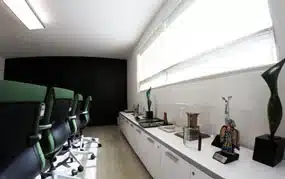 Una moderna sala de conferencias con sillas verdes, un largo armario blanco con varios elementos decorativos y grandes ventanales con las persianas cerradas.