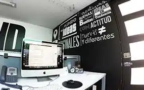 Una configuración de oficina moderna con computadora, teclado, mouse, teléfono y cámara. El muro muestra frases motivadoras en español.
