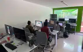 En una oficina luminosa de una agencia de publicidad y marketing, las personas trabajan diligentemente en escritorios de computadora, con varios monitores y sillas ergonómicas, todas ubicadas para aprovechar la luz natural que entra a través de una ventana grande.