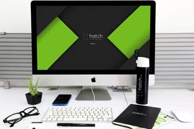 Una configuración de escritorio que incluye un iMac con un logotipo "hatch co" verde y negro en la pantalla, acompañado de una botella de agua, un cuaderno, un teclado, un mouse, una pequeña planta, anteojos y bolígrafos.