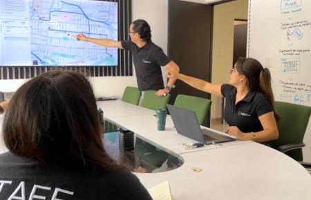 Dos personas con camisas negras señalan una pantalla grande que muestra un mapa durante una reunión de Servicios de Marketing en una sala de conferencias. Al fondo se ve una pizarra con notas.