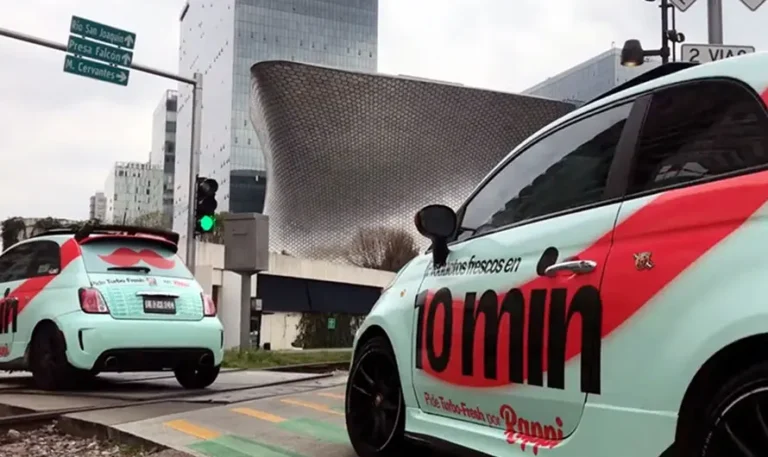 Dos pequeños autos con el texto "10 min" en el costado, parte de una activación de marca en CDMX, están estacionados cerca de una esquina en una zona urbana con arquitectura moderna.