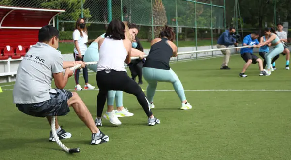 Dos equipos de personas participan en activaciones deportivas, jugando al tira y afloja en un campo de césped en la CDMX, tirando de una cuerda en direcciones opuestas. ¡Asegúrate de usar el calzado adecuado para un mejor agarre y rendimiento!