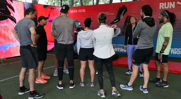 Un grupo de personas vestidas con ropa deportiva se encuentran en semicírculo en un interior, escuchando atentamente a un orador que analiza una estrategia de activación para nuevos productos deportivos frente a una pantalla grande.