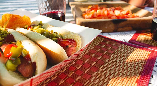 Fotografía de productos para contenido en redes: Dos hot dogs con salsa de tomate y salsa de tomate reposan en un plato junto a un vaso de bebida roja y un acompañamiento de tomates en rodajas, elegantemente dispuestos sobre una mesa con un tapete tejido rojo y beige.