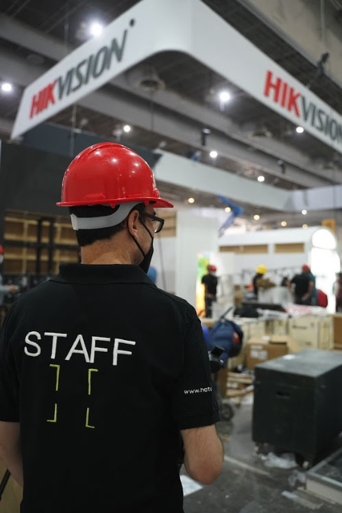 Un miembro del personal que lleva un casco rojo y una camisa negra con "STAFF" en la espalda se encuentra en un área de construcción interior debajo de un gran letrero que dice "HIKVISION", en representación de Hatch Co.