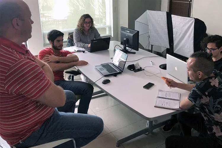 Cinco personas están sentadas alrededor de una mesa en una oficina, enfrascadas en una discusión. Algunos usan computadoras portátiles mientras que una persona tiene una libreta abierta. Al fondo, el equipo de fotografía deja entrever los proyectos creativos que se llevan a cabo en esta agencia de diseño.