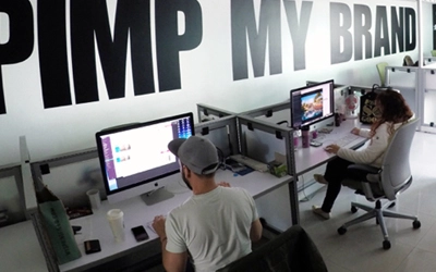 Dos personas trabajando en escritorios con computadoras en un ambiente de oficina. El texto grande en la pared detrás de ellos dice "PROTUNO MI MARCA". Esta bulliciosa escena es parte de Hatch Co, una dinámica agencia de marketing que está causando sensación en la industria.