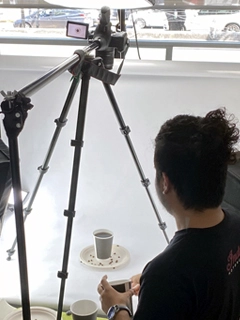 Una persona está detrás de una cámara montada sobre un trípode, fotografiando una taza sobre un platillo con algunos frijoles esparcidos en un estudio bien iluminado, capturando a la perfección la esencia de un producto elegante fotografiado para una agencia de marketing.