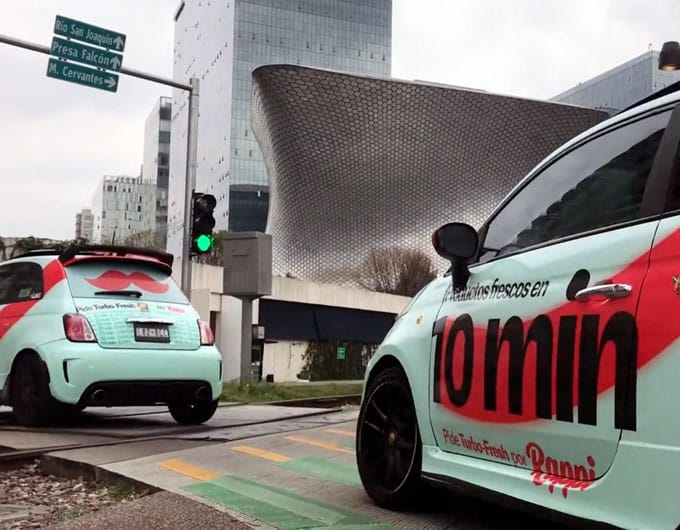 Dos coches de color turquesa con bigotes rosas y la marca del servicio de reparto están detenidos en un semáforo en una zona urbana con edificios modernos al fondo, ejemplificando un llamativo marketing callejero.