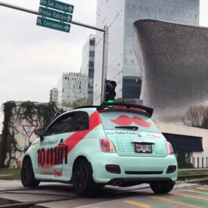 Un automóvil compacto de color azul claro y rojo con un equipo sensor verde en el techo circula por una calle en una zona urbana cerca de edificios altos y señales de tráfico.