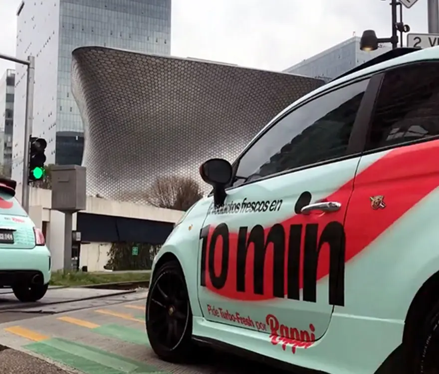 Dos coches con anuncios de un servicio de reparto se encuentran en un semáforo, con un edificio moderno como fondo. El anuncio en el coche dice "Refrescos fríos en 10 min", mostrando una marca innovadora y activaciones en México.