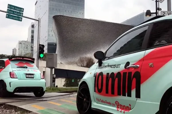 Dos pequeños autos con el texto "10 min" en el costado, parte de una activación de marca en CDMX, están estacionados cerca de una esquina en una zona urbana con arquitectura moderna.