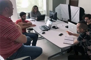 Cinco personas están sentadas alrededor de una mesa con computadoras portátiles y cuadernos, enfrascadas en una discusión seria. En el fondo hay una pizarra y reflectores de luz, lo que indica que hay una agencia de marketing CDMX en plena actividad colaborando en nuevas estrategias.