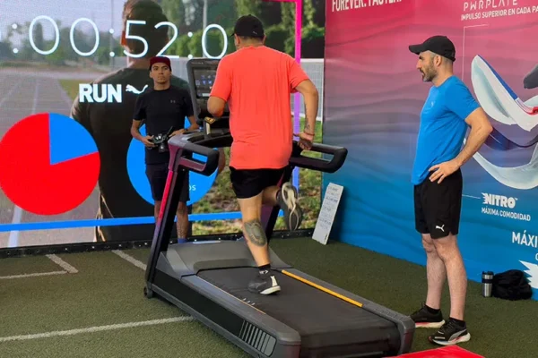 Un hombre corre en una cinta de correr en un recinto cerrado, completamente equipado con ropa deportiva para una activación de marca deportiva. Otros dos hombres están de pie cerca, observando mientras una gran pantalla muestra un cronómetro y un gráfico circular.
