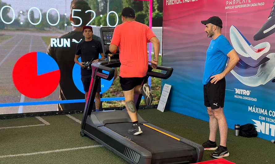 Un hombre corre en una cinta de correr en un recinto cerrado, completamente equipado con ropa deportiva para una activación de marca deportiva. Otros dos hombres están de pie cerca, observando mientras una gran pantalla muestra un cronómetro y un gráfico circular.